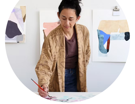 Artist painting abstract artwork at a table, with finished pieces displayed on the wall behind.