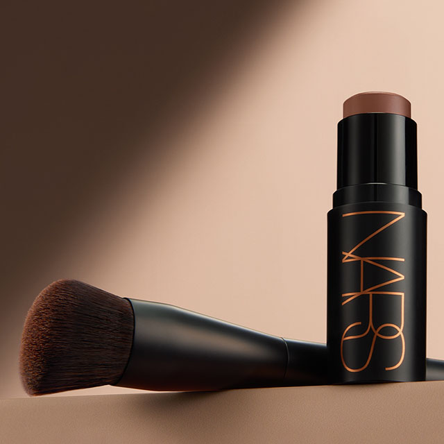 A makeup brush and a NARS foundation stick against a beige background.