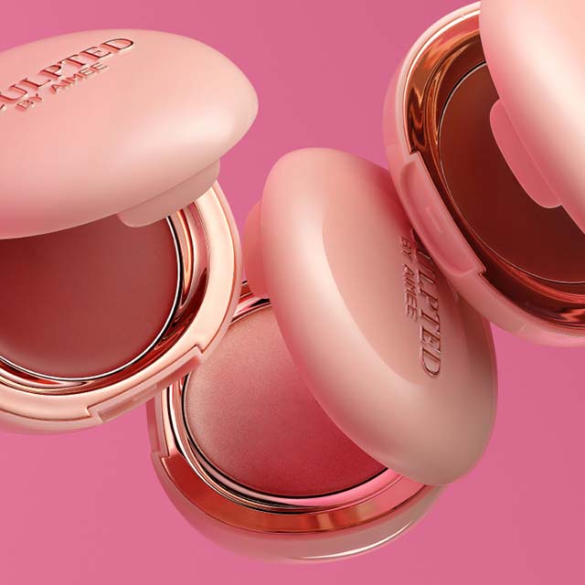 Three pink and gold Sculpted with Aimee makeup compacts with mirror reflections on a pink background.