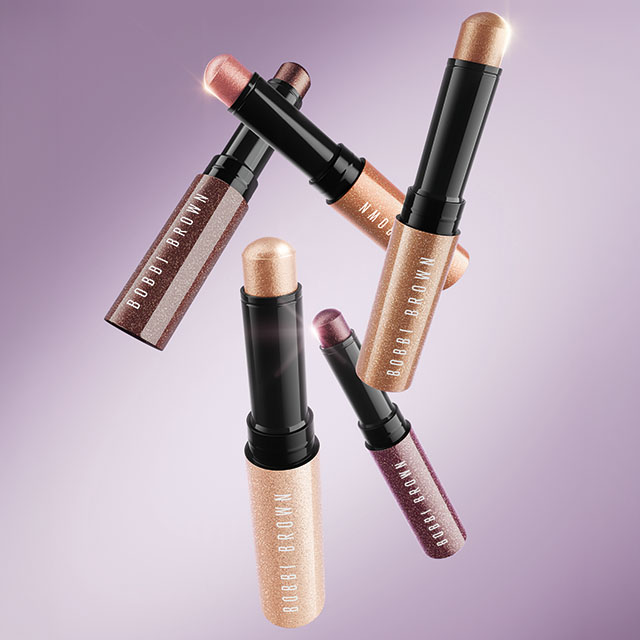 Five glittery Bobbi Brown makeup sticks in various shades against a purple background.
