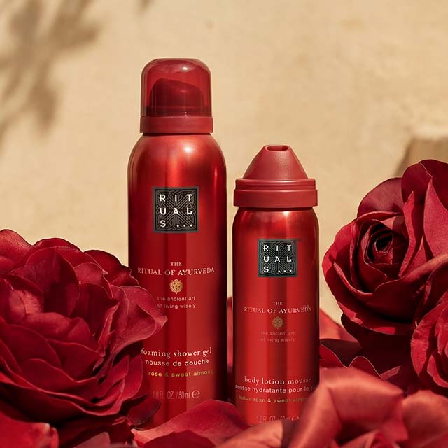Two red Rituals beauty products surrounded by red roses against a beige background.