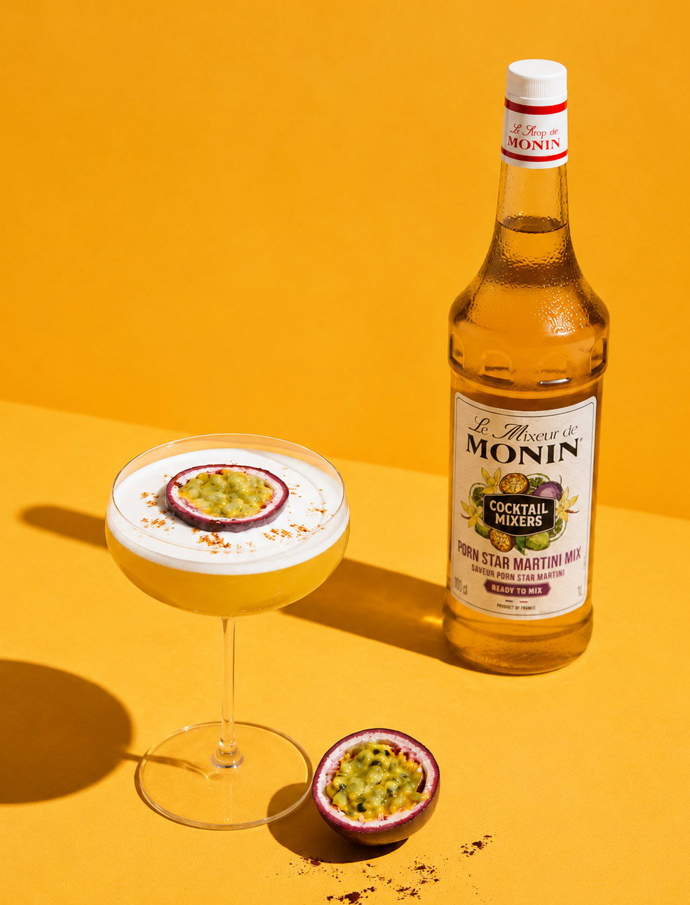 Passion fruit martini in coupe glass garnished with halved passion fruit and Monin martini mixer bottle on yellow background