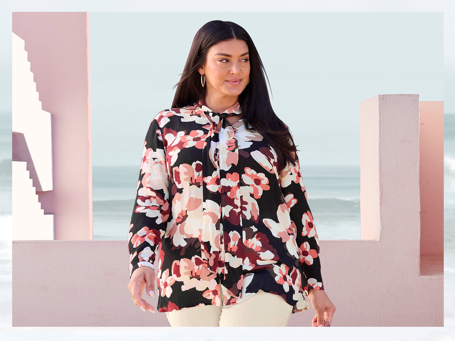 Ein Model trägt eine langärmelige Bluse mit großem, buntem Blumenmuster und helle Hose. Im Hintergrund Architektur in Rosa und das Meer.