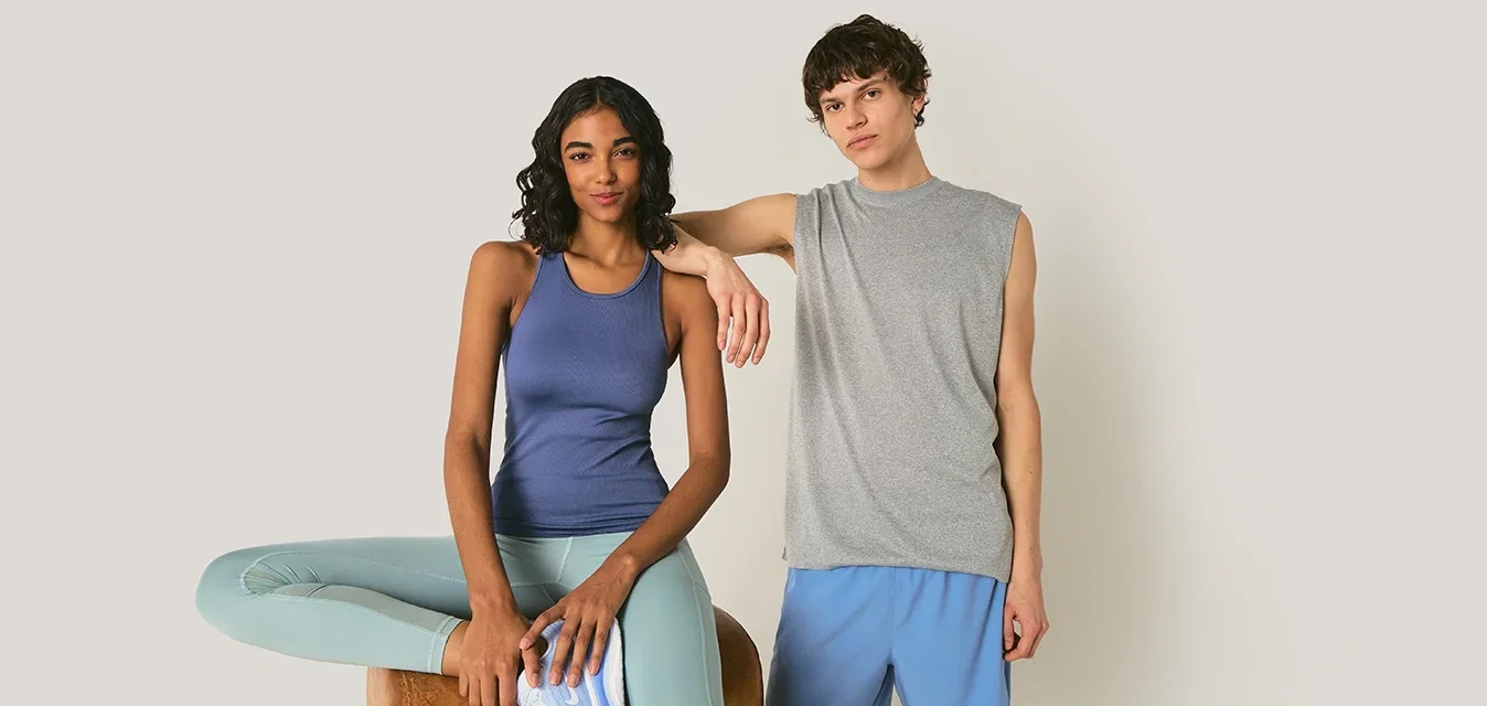 Dos adultos jóvenes con ropa deportiva informal; uno sentado en un taburete de madera con una pierna doblada, y el otro de pie apoyando un brazo en el hombro de la persona sentada.