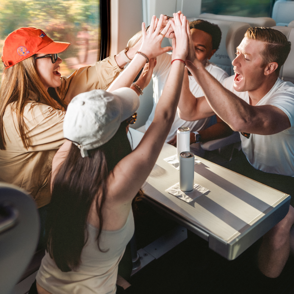 A group of young adults sit around a table on a moving train, raising their hands together in a celebratory toast.