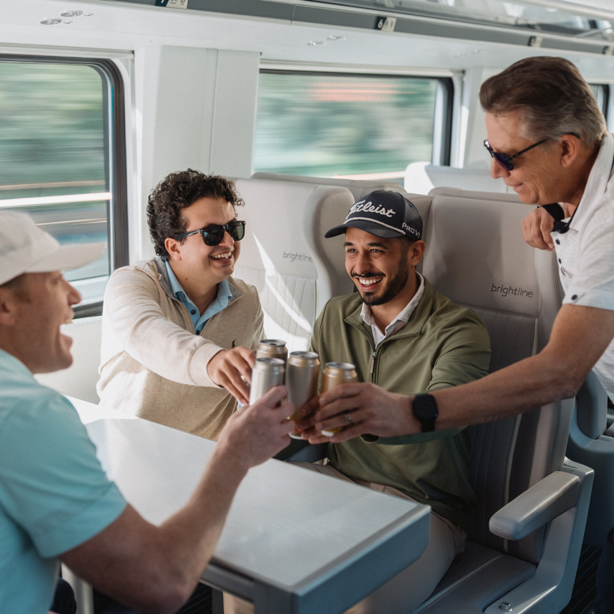 A group of adults sit around a table on a modern passenger train, clinking canned drinks together in a toast.