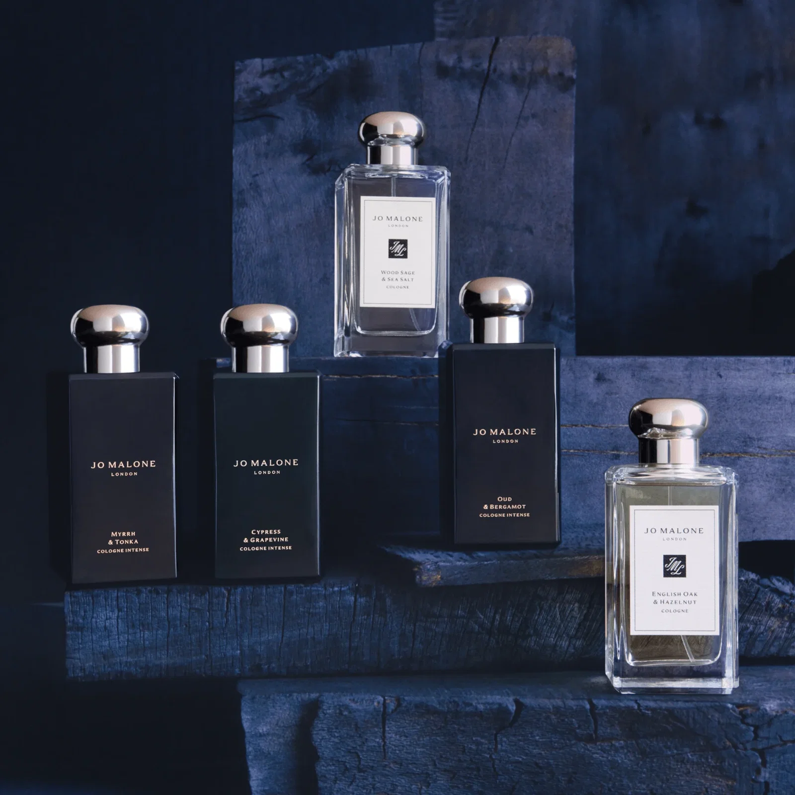 Five Jo Malone men's cologne bottles arranged on chunks of blue wood against a dark blue background.