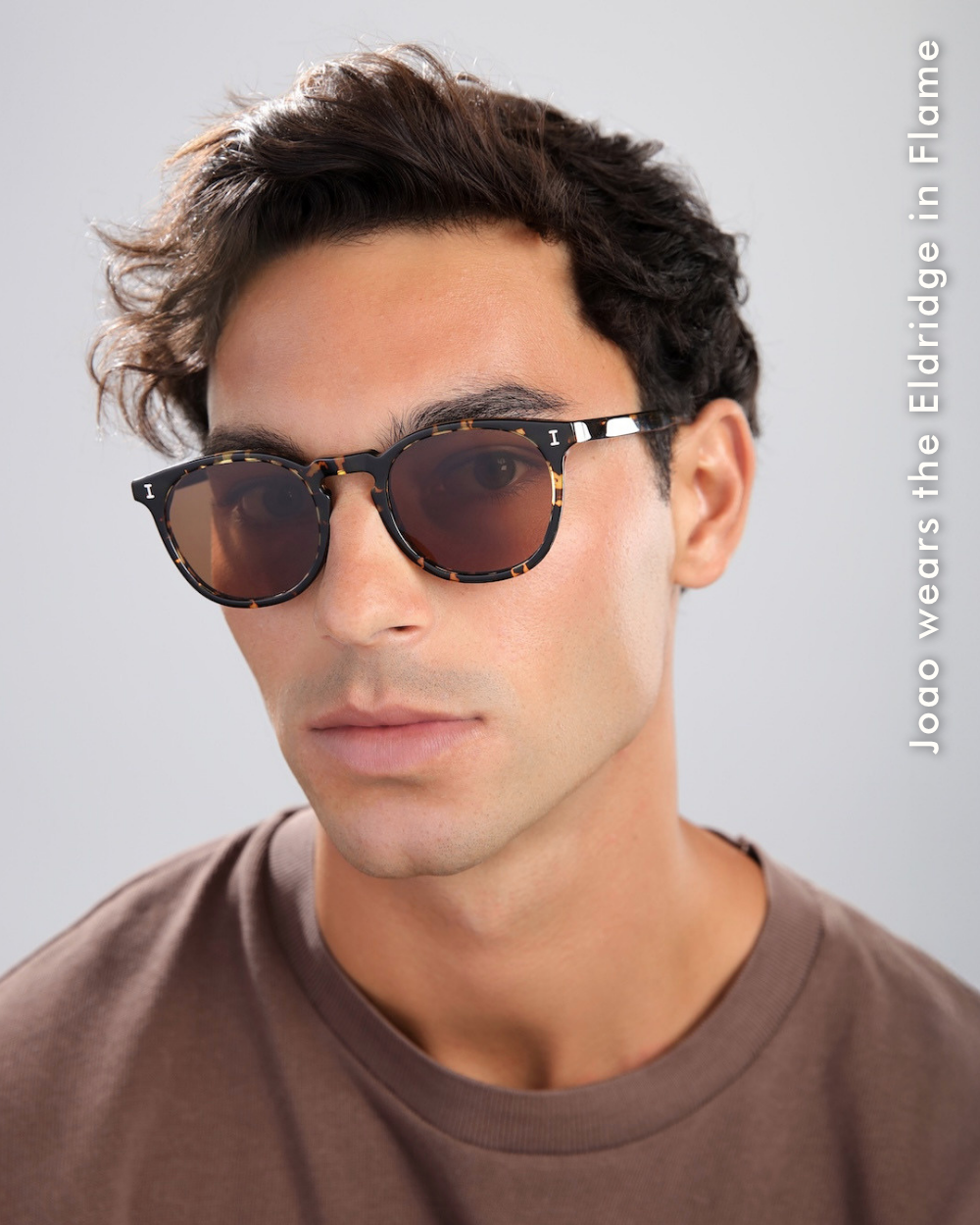 A portrait of a man named Joao wearing the Eldridge in Flame, which features a dark tortoise-shell patterned frame with warm brown-tinted lenses.