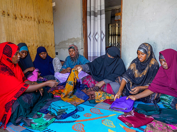a group of 7 women sit in a semi-circle on a mat, reviewing colourfully dyed dresses.
