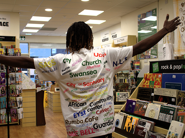 A man stood in a bookshop, facing away from the camera. The back of his t-shirt is covered in words, including “community,” “city of sanctuary,” “Swansea.”