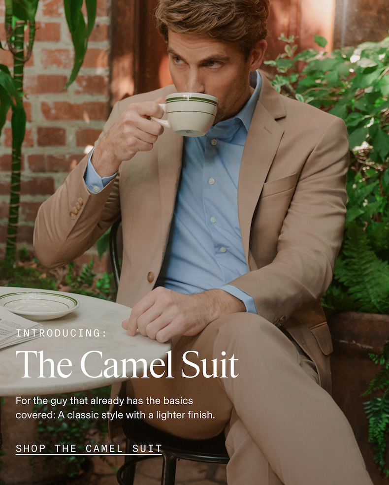 Man in a camel suit and light blue dress shirt seated at a marble bistro table in a lush garden courtyard, sipping from a ceramic teacup. Overlaid text reads: INTRODUCING: The Camel Suit. For the guy that already has the basics covered: A classic style with a lighter finish. SHOP THE CAMEL SUIT