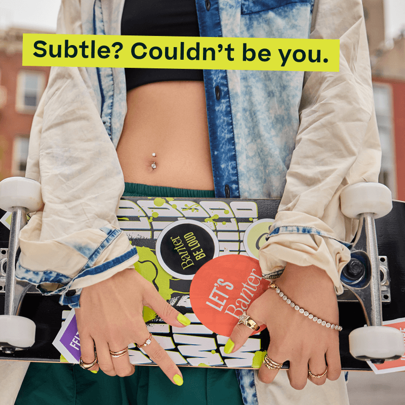 Model holding sticker-covered skateboard with double-gem belly piercing, gold rings & tennis bracelet. Subtle? Not you.