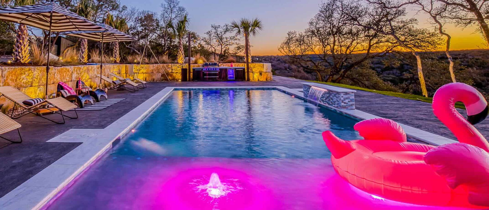 Luxury modern vacation rental in Austin, TX with resort-style backyard, glowing swimming pool, covered patio, outdoor lounge seating, and contemporary multi-story architecture at sunset.