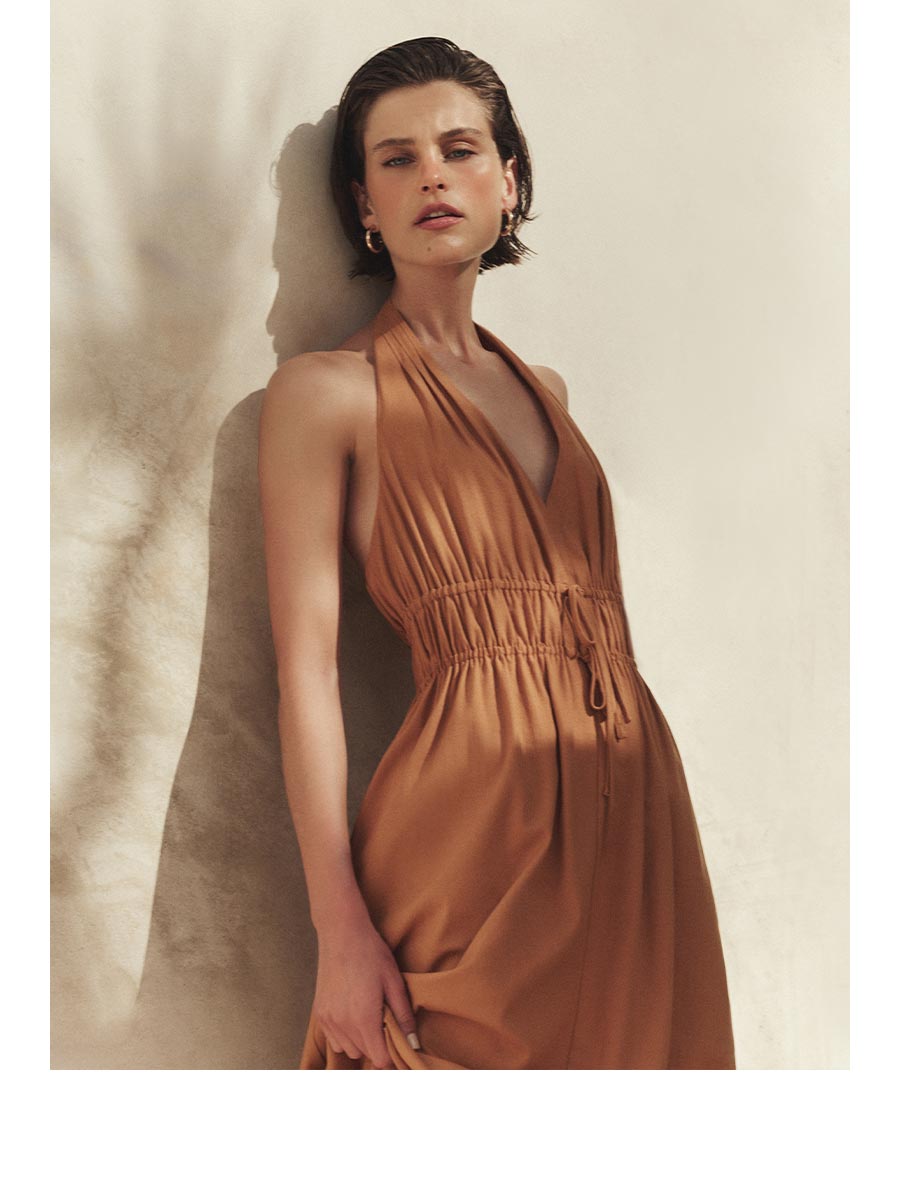 Image of a woman wearing an earthy orange linen dress leaning against a beige wall with palm shadows.