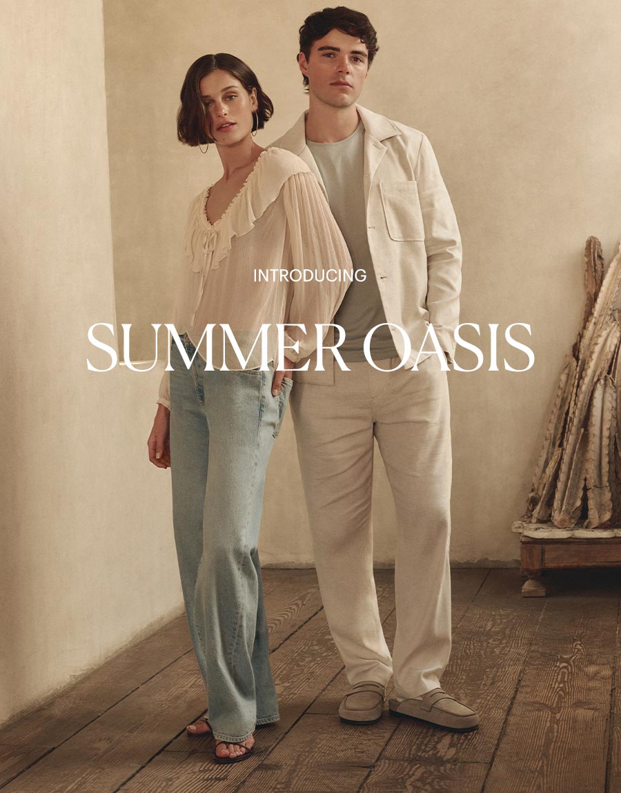 Introducing Summer Oasis - Featuring a woman wearing a light cream colored silk flowy blouse with wide leg light blue wash denim jeans and black strappy sandals standing next to a man wearing a white linen jacket, matching linen pants, a light blue t-shirt and light tan suede loafers all by PAIGE.