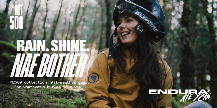 Smiling cyclist in helmet and yellow waterproof MT500 jacket in woodland, promotional text 'Rain. Shine. NAE BOTHER' with Endura logo