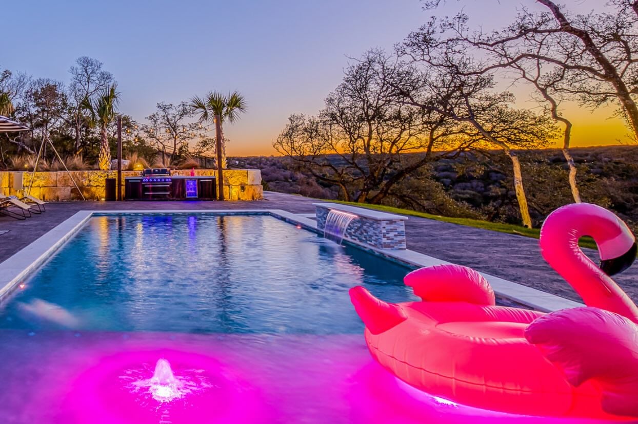 Luxury modern vacation rental in Austin, TX with resort-style backyard, glowing swimming pool, covered patio, outdoor lounge seating, and contemporary multi-story architecture at sunset.