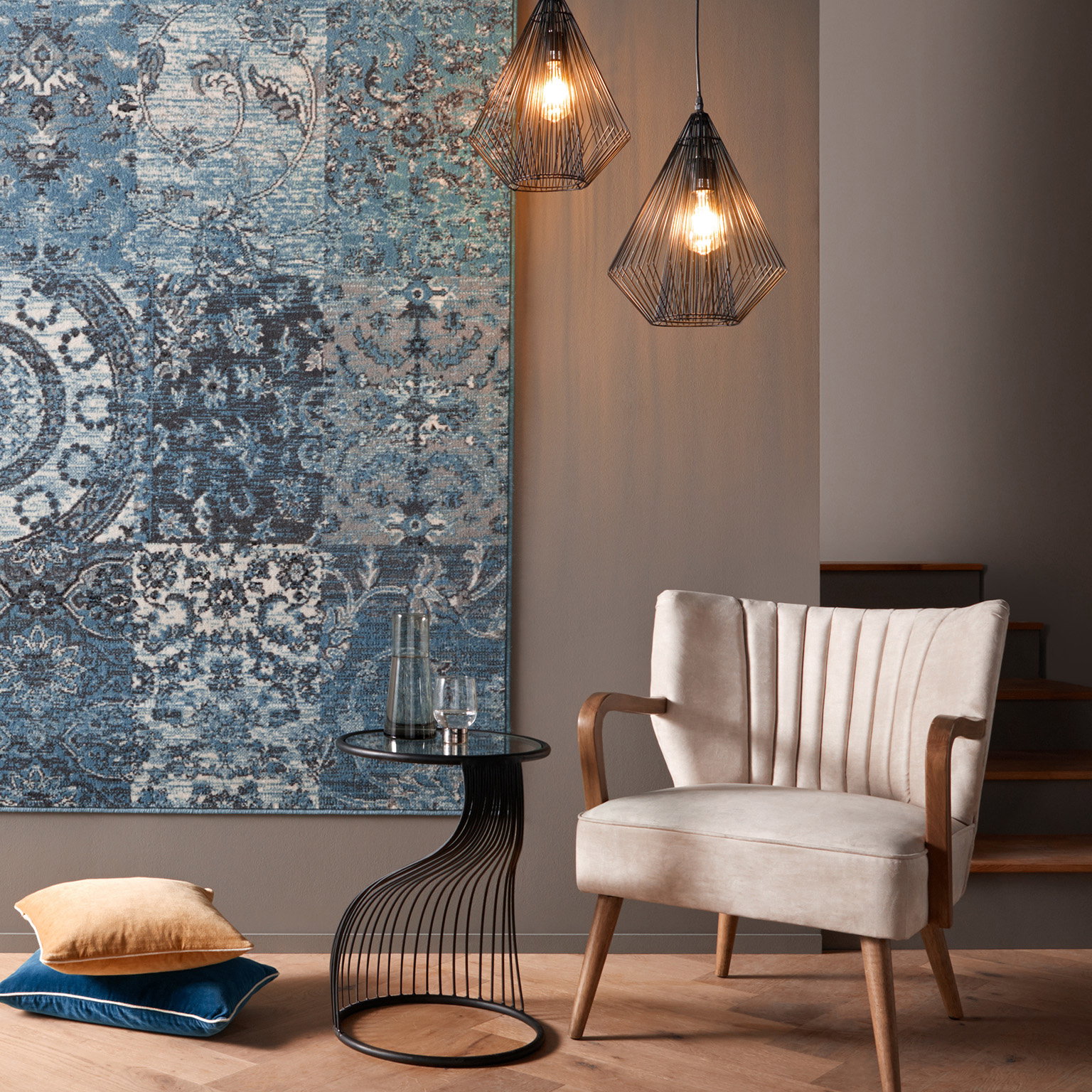 Gezellige zithoek met beige stoel, kleine zwarte tafel, twee kussens op de vloer en blauw patroon wandkleed.