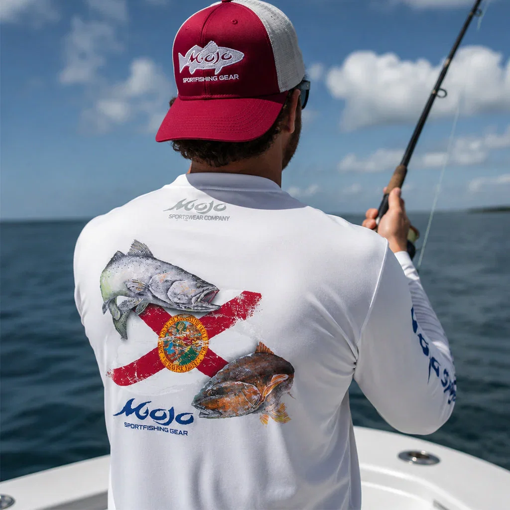 Fisherman wearing Mojo Sportswear red cap and white long-sleeve shirt with Florida flag and fish design while holding fishing rod on boat.