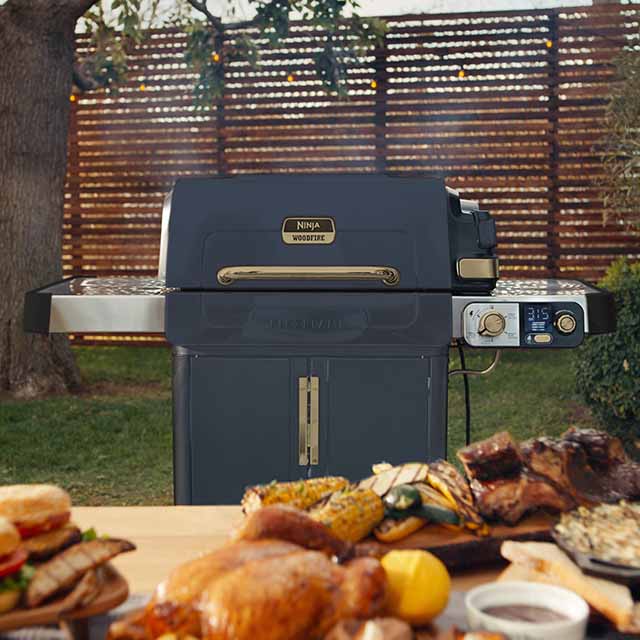 A grill in a backyard setting with various cooked foods on a table in the foreground.