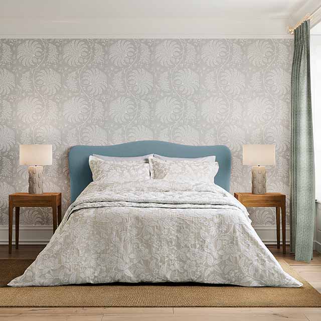 A bed with a light blue headboard, white bedding, and two bedside tables with lamps. The room has patterned wallpaper and a window with a curtain.