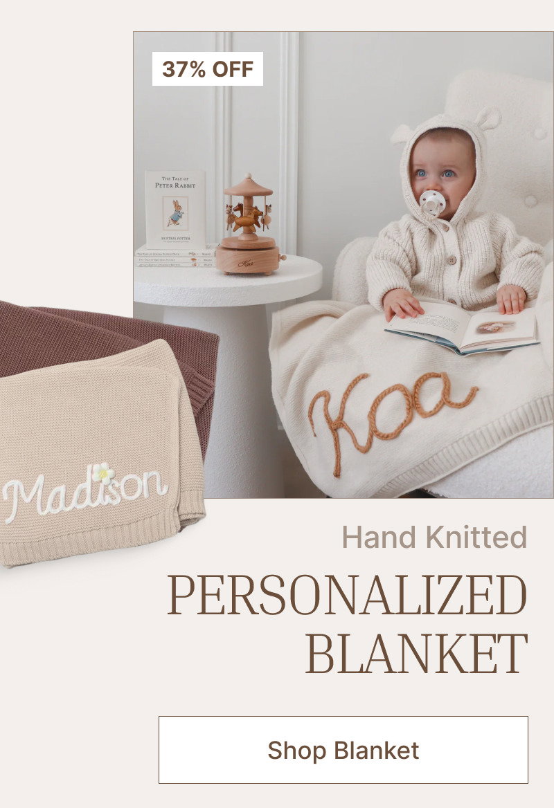 Baby sitting on a bed near a personalized knitted blanket, wooden toy decor, and soft nursery accessories.