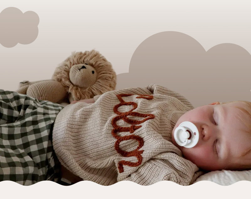 Baby sleeping beside a knitted teddy bear on a soft blanket with neutral cloud shapes in the background.