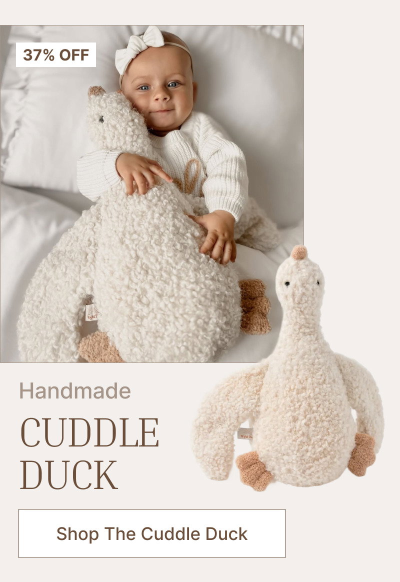 Baby in a white outfit lying beside a large cream plush duck toy on a light bed with soft neutral styling.
