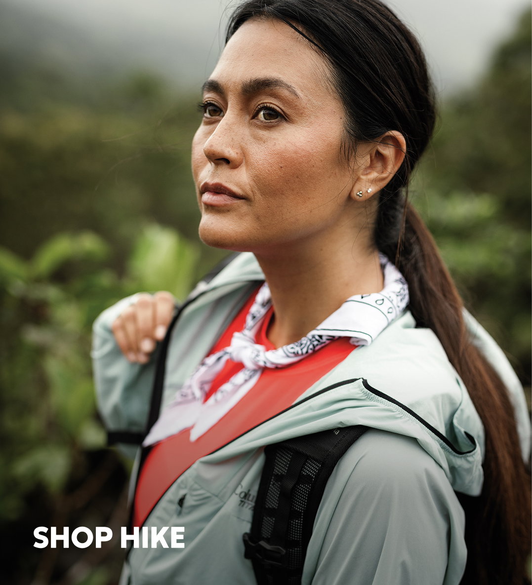 A woman wearing a red shirt, light green windbreaker, and purple neckerchief, standing in a green forest with text: SHOP HIKE