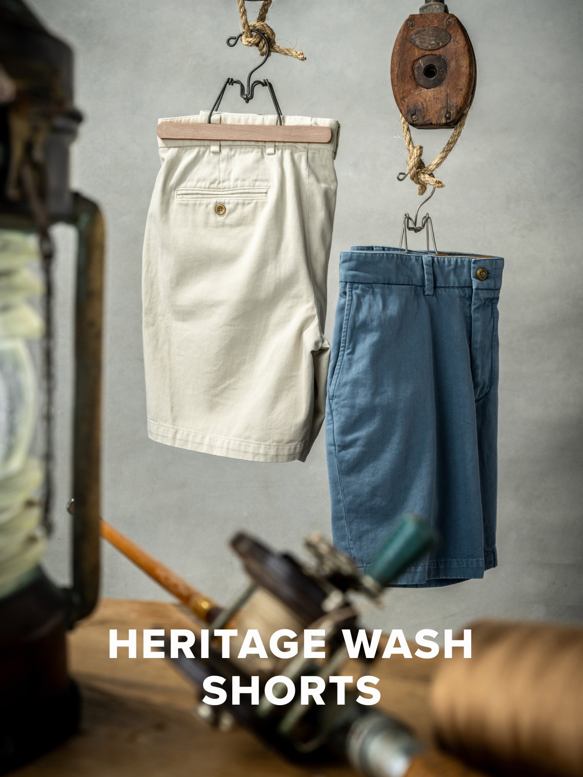 A photo of Bills Khakis new Bone and Dusty Blue Heritage Wash Shorts hanging from industrial pulleys against a grey backdrop. Vintage mariner items sit out of focus in the foreground