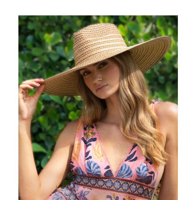 A woman in a wide-brimmed straw hat poses outdoors, wearing a floral dress.