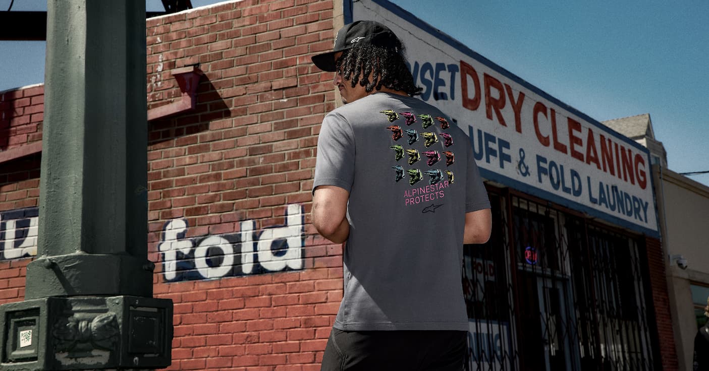 Man in a grey t-shirt with a graphic of helmets and text "ALPINESTAR PROTECTS".