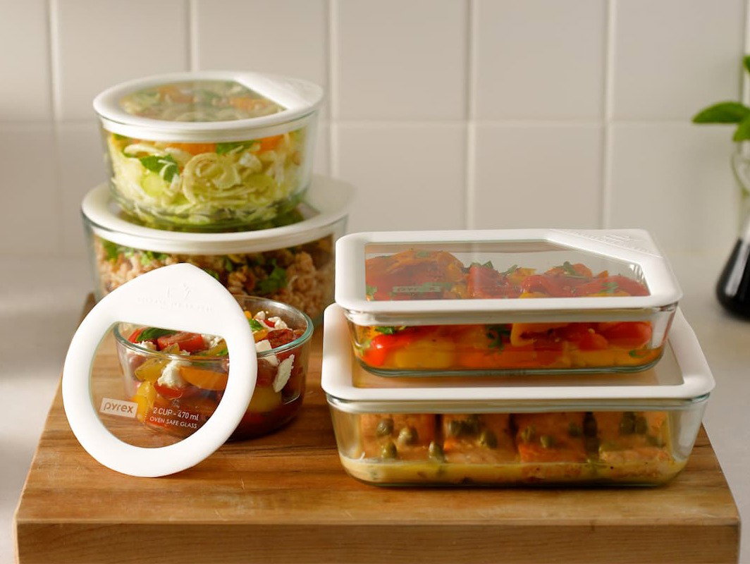 Five stacked Pyrex® glass containers in round and rectangular shapes, one open with food inside, showing clear lids and storage design.