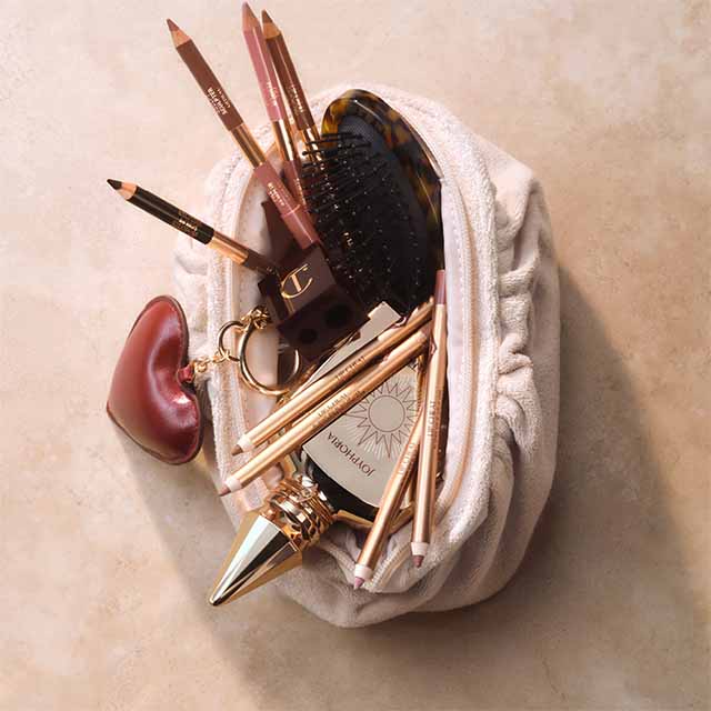 A makeup bag containing beauty products, pencils, a compact mirror, and a hairbrush on a beige marble surface.
