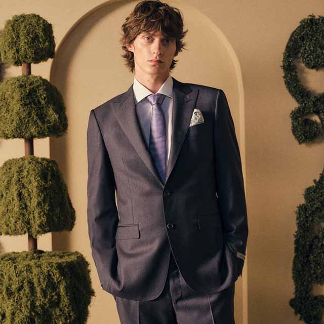 A person in a suit stands in a sophisticated setting with topiary plants.