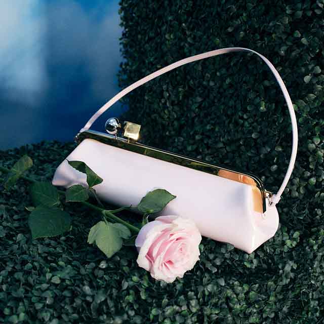 A pink handbag with a strap rests on greenery next to a pink rose.