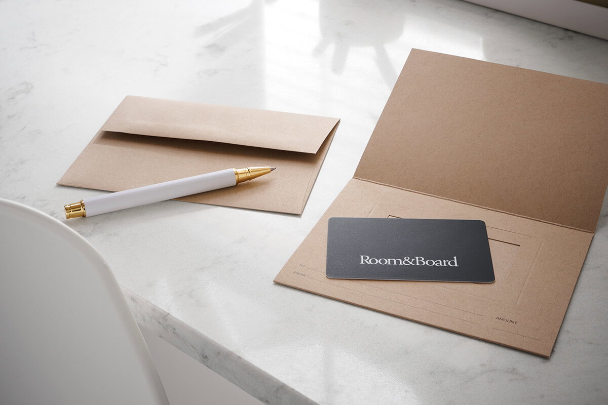 Room&Board gift card placed on a kraft card holder beside a matching envelope and white pen on a marble surface.