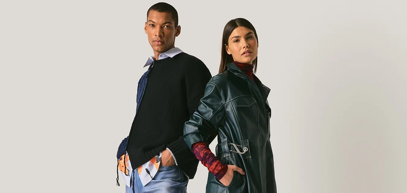 Hombre y mujer de pie espalda con espalda, vistiendo atuendos modernos en capas con mezclilla, gabardina de cuero y zapatos casuales sobre fondo neutro.