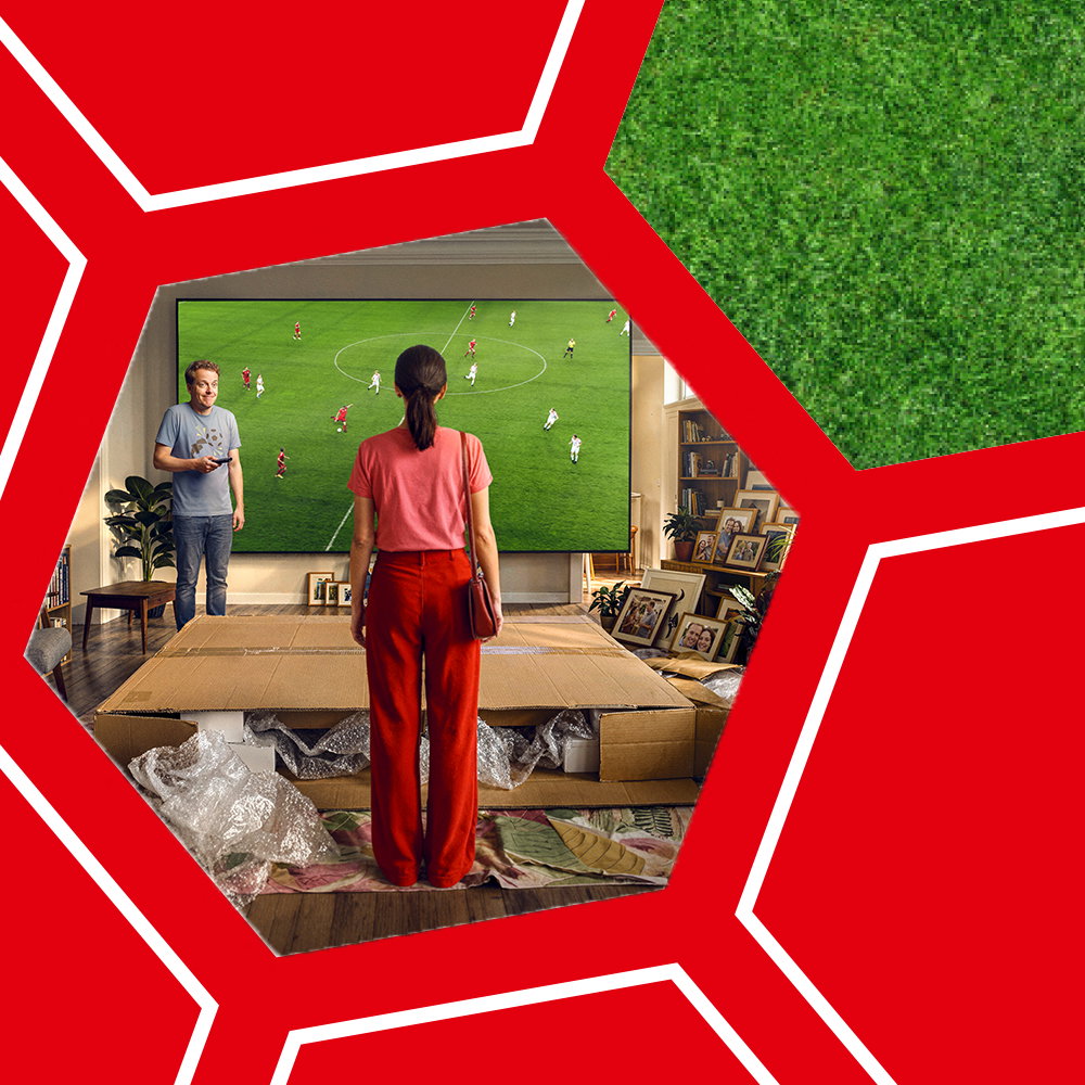 Zwei Personen stehen vor einem großen Fernseher, auf dem ein Fußballspiel gezeigt wird, umgeben von roten Fünfecken und grünem Rasenmuster