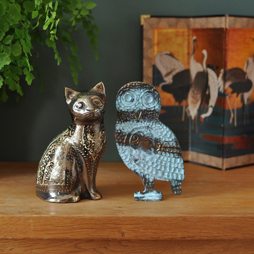 Engraved brass cat ornament and bronze owl figure displayed on a wooden sideboard. - Shop Replicas and Ornaments.