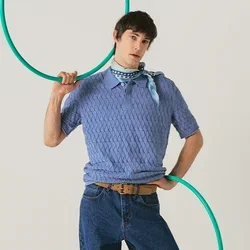 Persona con maglione blu e jeans posa con un hula hoop verde, in piedi davanti a uno sfondo chiaro e uniforme.