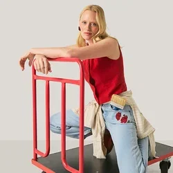 Frau in rotem Oberteil und Jeans kniet auf einem schwarzen Plattformwagen mit rotem Griff vor einfachem Hintergrund.