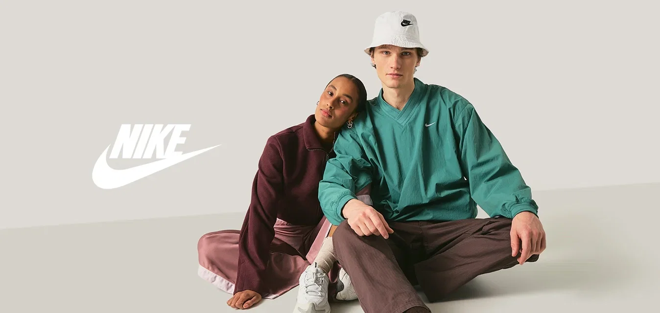 Zwei junge Erwachsene sitzen auf dem Boden, der eine lehnt seinen Kopf an die Schulter des anderen, beide tragen legere sportliche Kleidung und Sneakers.
