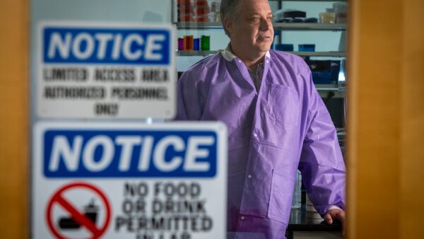 A man in a purple lab coat walks by a door with “notice” signs on it that the area is for authorized personnel only and no food permitted in the lab. 