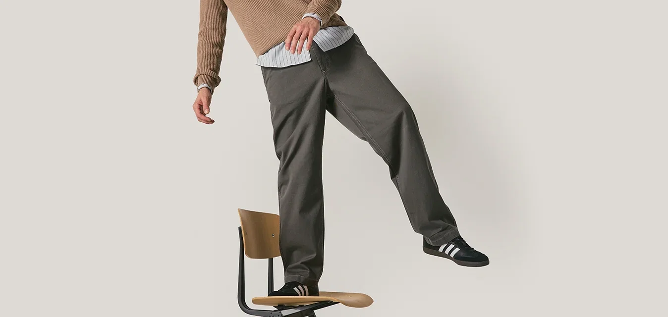 Pantalon gris à coupe décontractée, avec des coutures détaillées, porté avec un pull texturé beige et des chaussures noires avec des rayures blanches. Debout sur une chaise.
