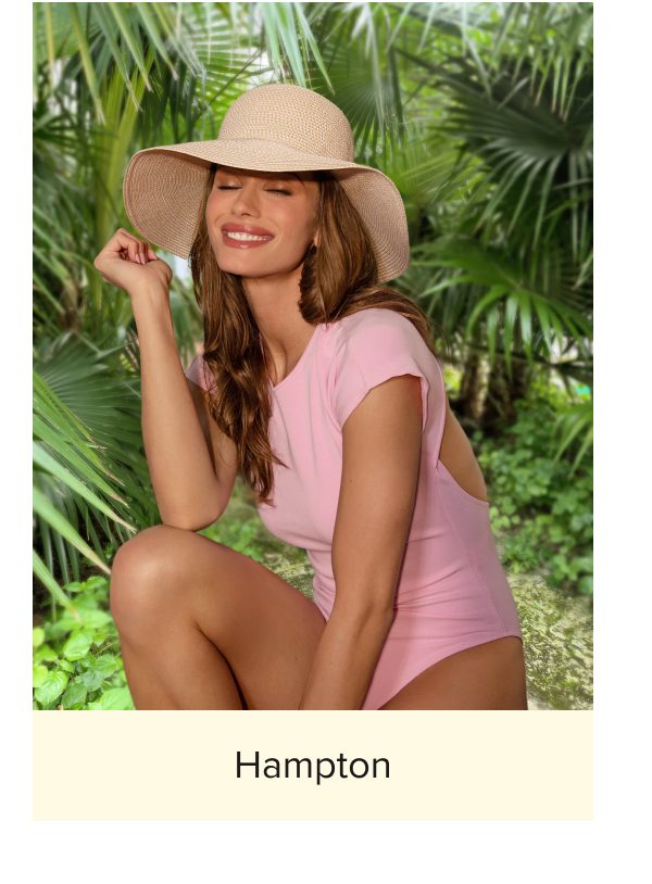 Woman in a pink swimsuit and wide-brimmed hat, smiling in a tropical setting.