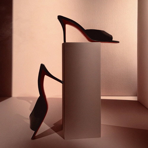 Two black high-heeled shoes with red soles are artistically arranged in a minimalist setting. One shoe is upright on a pedestal, while the other rests against it, creating an elegant, symmetrical composition. The background is softly lit with warm tones, enhancing the luxurious feel.