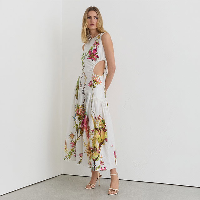A woman in a floral cutout dress and heels stands in a minimalist corner.