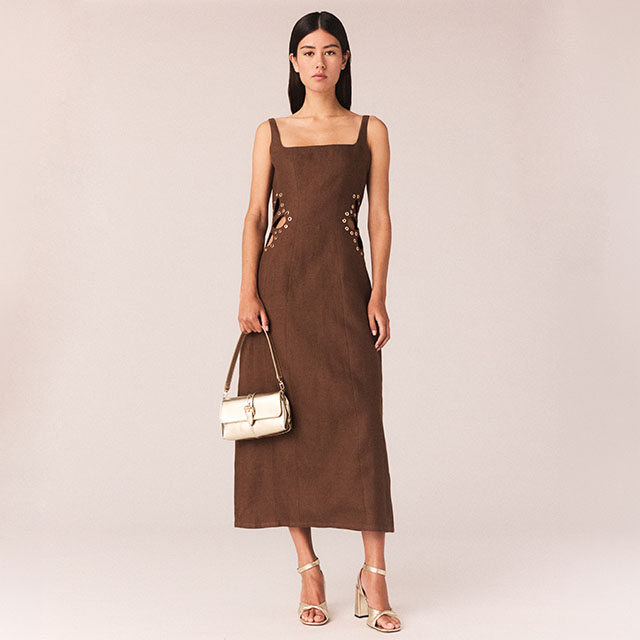 A woman wearing a sleeveless, brown, ankle-length dress with decorative floral embroidery near the hips. She holds a small metallic handbag in her left hand and is wearing strappy high-heeled sandals. The background is plain and neutral. 