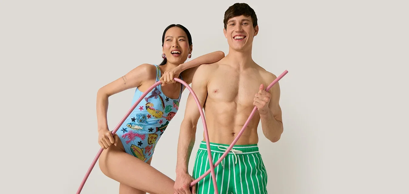 Lachende vrouw in een badpak met patroon en man in groene gestreepte zwembroek houden roze hula hoops vast tegen een effen achtergrond.